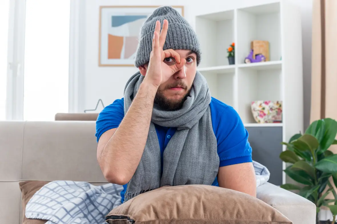 impressed young freezing man wearing scarf winter hat sitting sofa living